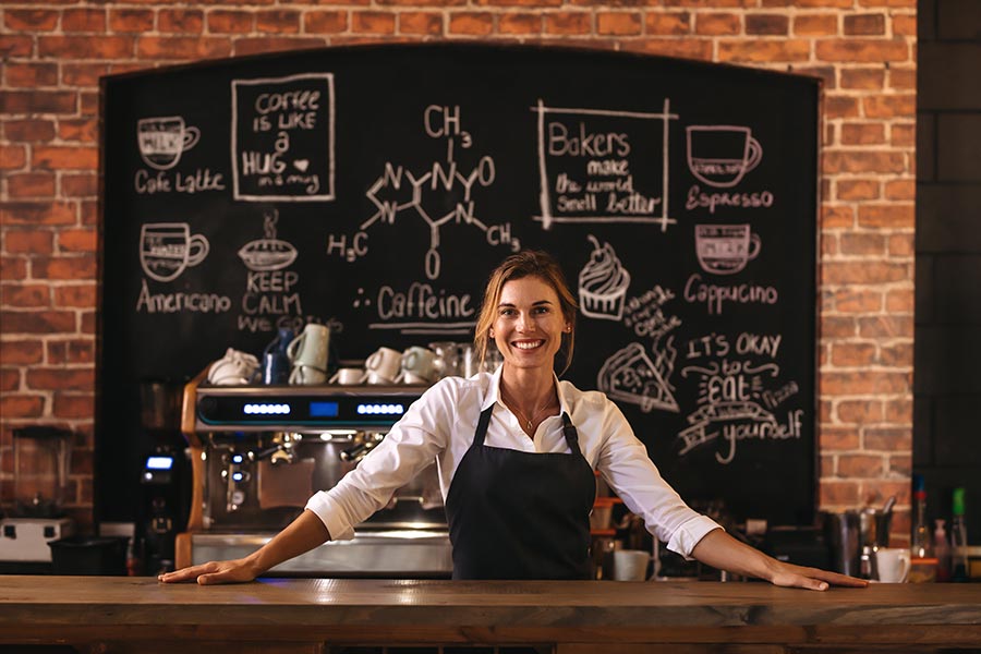 Business Insurance - Cafe Owner Stands Behind the Counter, Brick Wall and Chalkboard Menu Behind Her